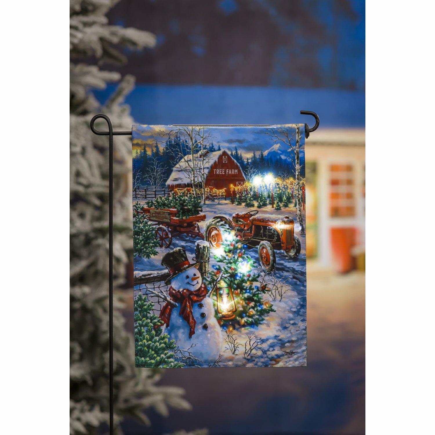 The Tree Farm Solar LED garden flag features a snowy Christmas tree farm complete with a tractor, wagon, barn, and snowman.