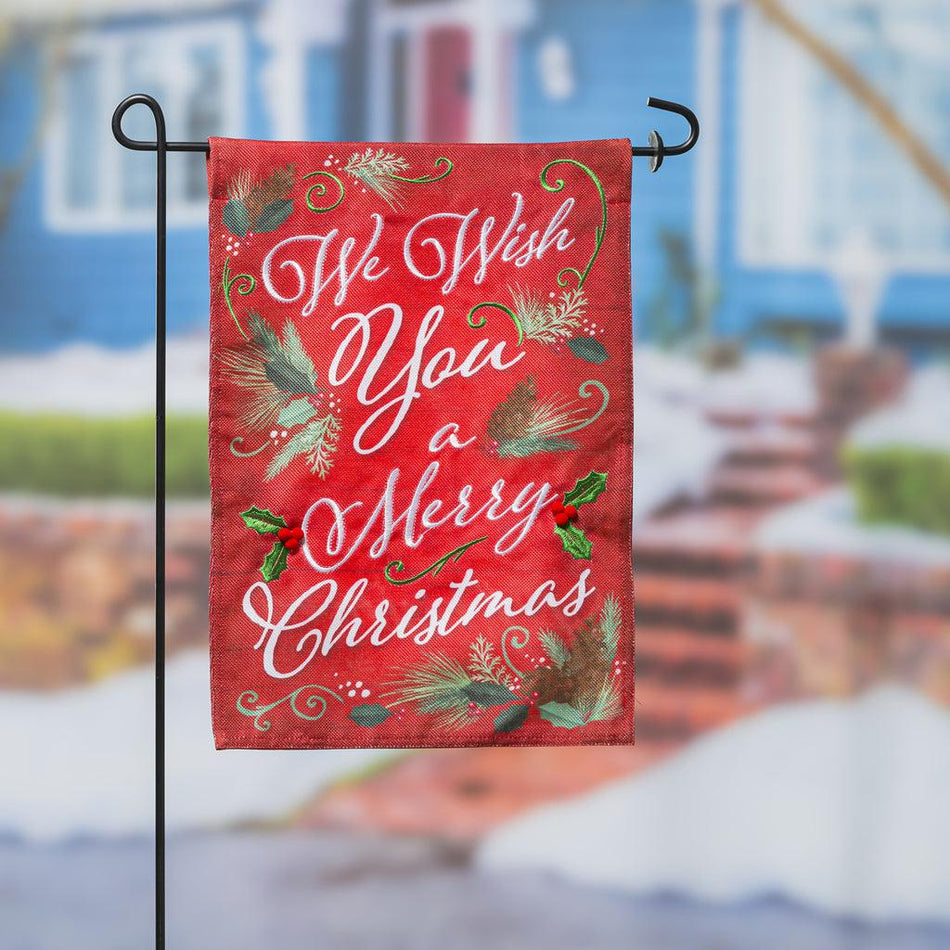 The We Wish You A Merry Christmas garden flag features the words "We Wish You A Merry Christmas" accented by pine and holly on a bright red background.