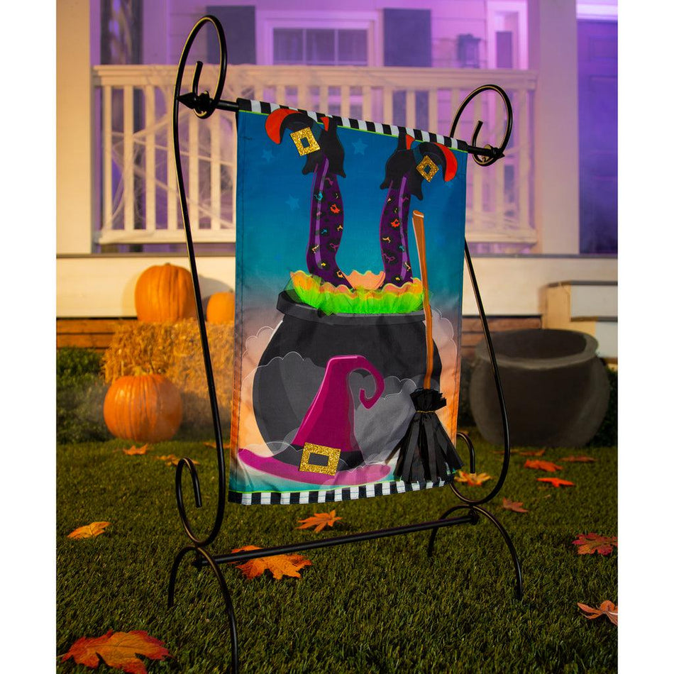 The Witches Cauldron garden flag features a witches legs sticking out of a cauldron and her hat and broom leaning against the cauldron.