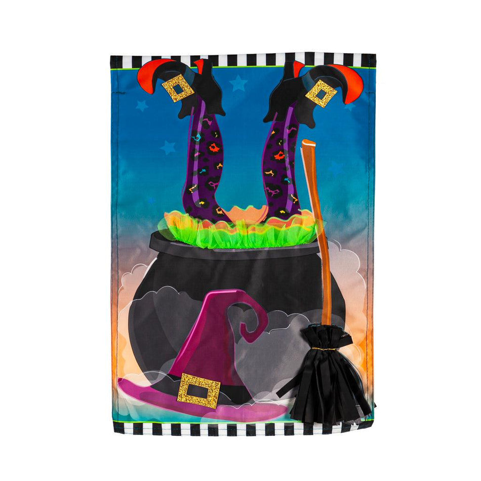 The Witches Cauldron garden flag features a witches legs sticking out of a cauldron and her hat and broom leaning against the cauldron.