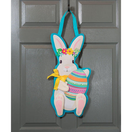 The Easter Bunny Door Décor features a white bunny with a floral crown and yellow ribbon holding an Easter egg. 