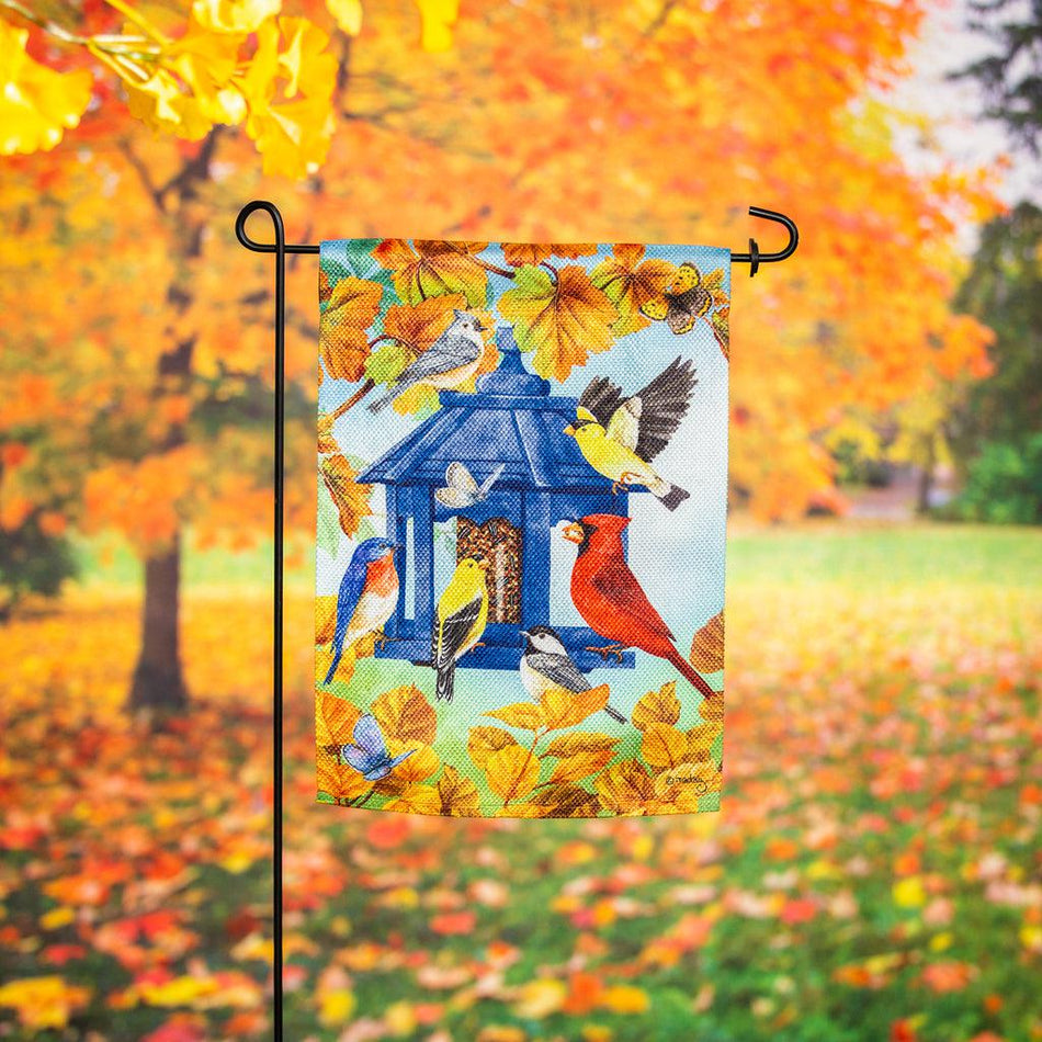 The Fall Feeder garden flag features a multitude of birds feasting from a blue bird feeder surrounded by fall leaves.
