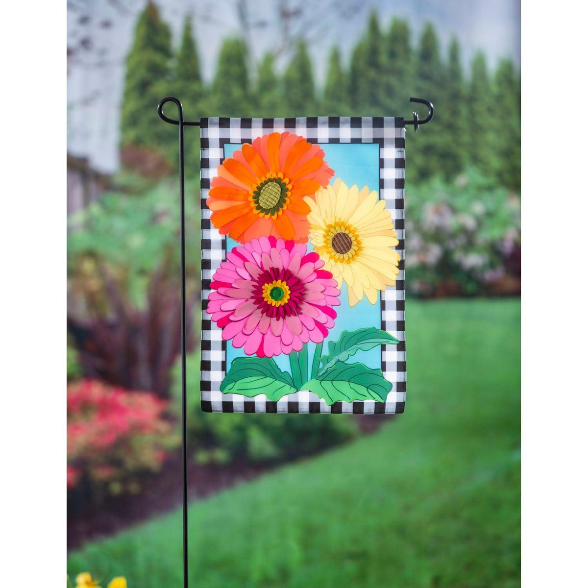 The Gerbera Daisy Trio garden flag features an orange, a yellow, and a pink Gerbera Daisy surrounded by a black and white checked border.
