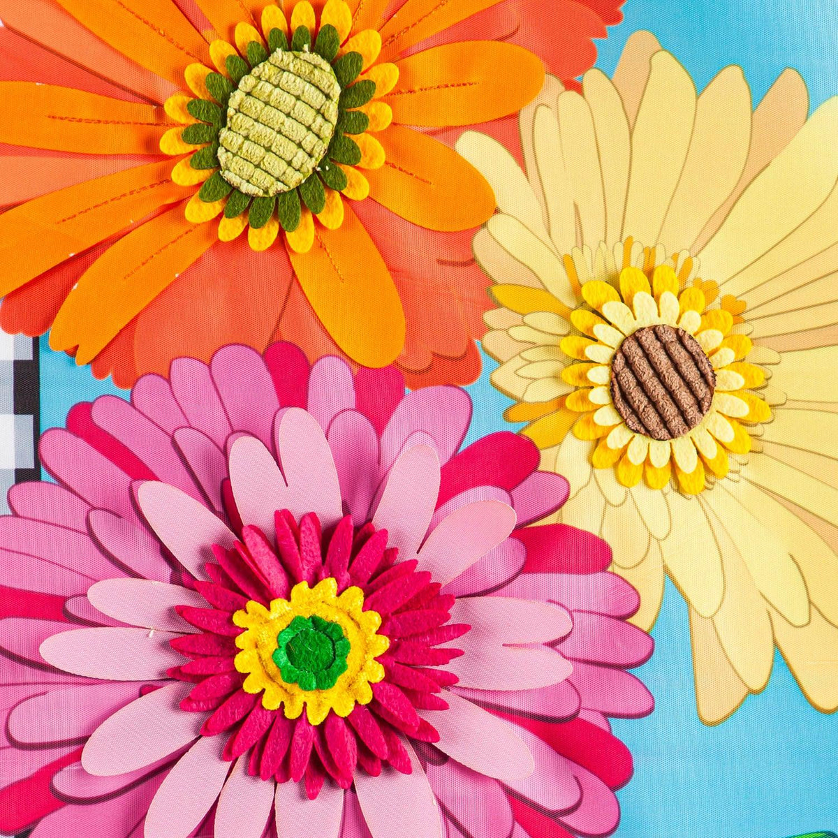 The Gerbera Daisy Trio garden flag features an orange, a yellow, and a pink Gerbera Daisy surrounded by a black and white checked border.