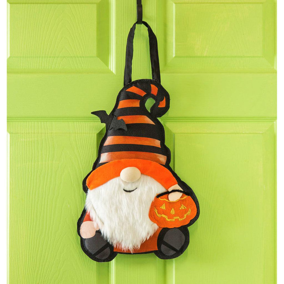 The Halloween Gnome door décor features a smiling gnome dressed in orange and black stripes and holding a jack-o-lantern bowl.