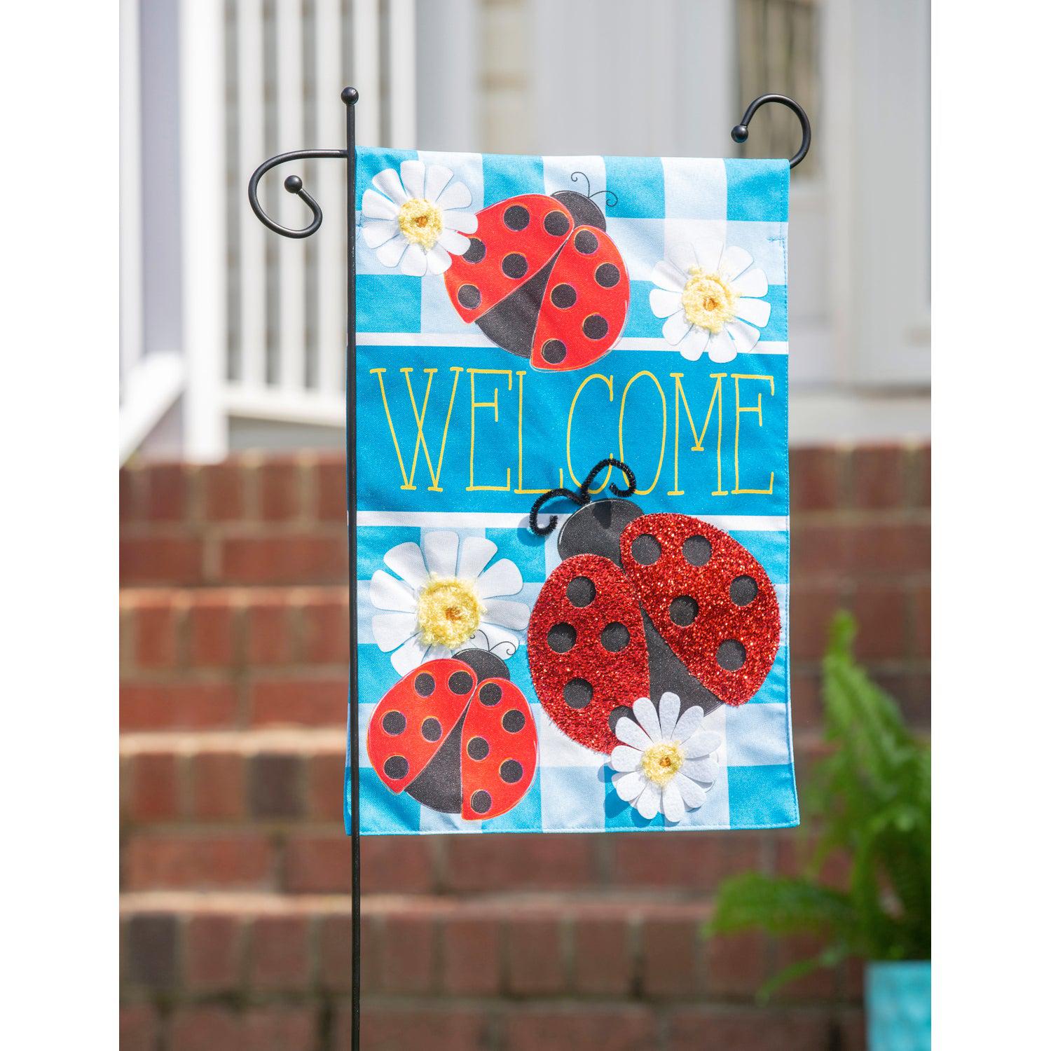 The Ladybug Plaid Welcome garden flag features a trio of ladybugs on a bright blue checked background with white daisies and the word "Welcome".