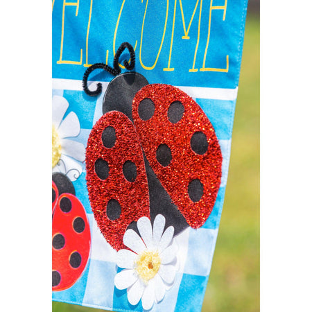 The Ladybug Plaid Welcome garden flag features a trio of ladybugs on a bright blue checked background with white daisies and the word "Welcome".