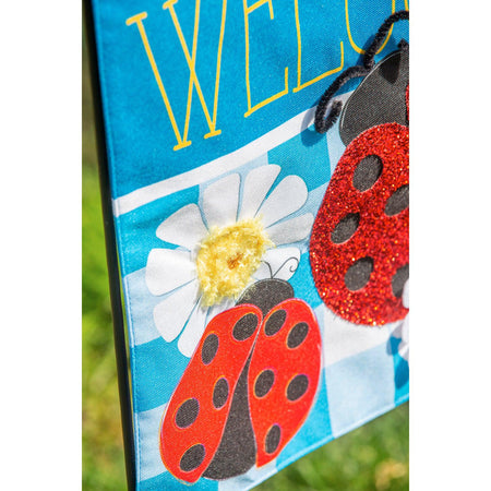 The Ladybug Plaid Welcome garden flag features a trio of ladybugs on a bright blue checked background with white daisies and the word "Welcome".