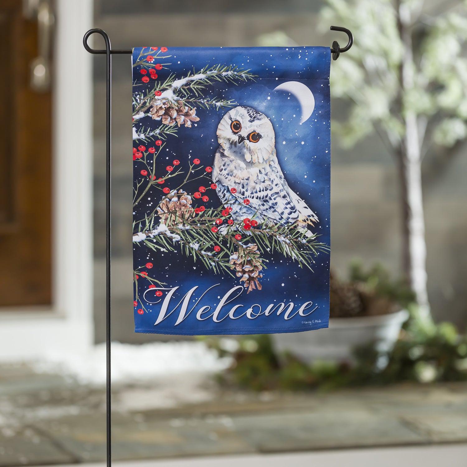 The Owl's Winter Greeting garden flag features a snowy owl at night resting on a pine branch along with the word "Welcome". 