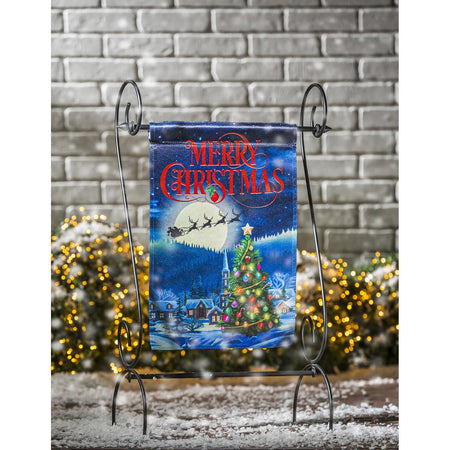 The Santa's Sleigh on Christmas garden flag features Santa and his reindeer flying over a small village with a beautiful Christmas tree, and the words "Merry Christmas" across the top. 