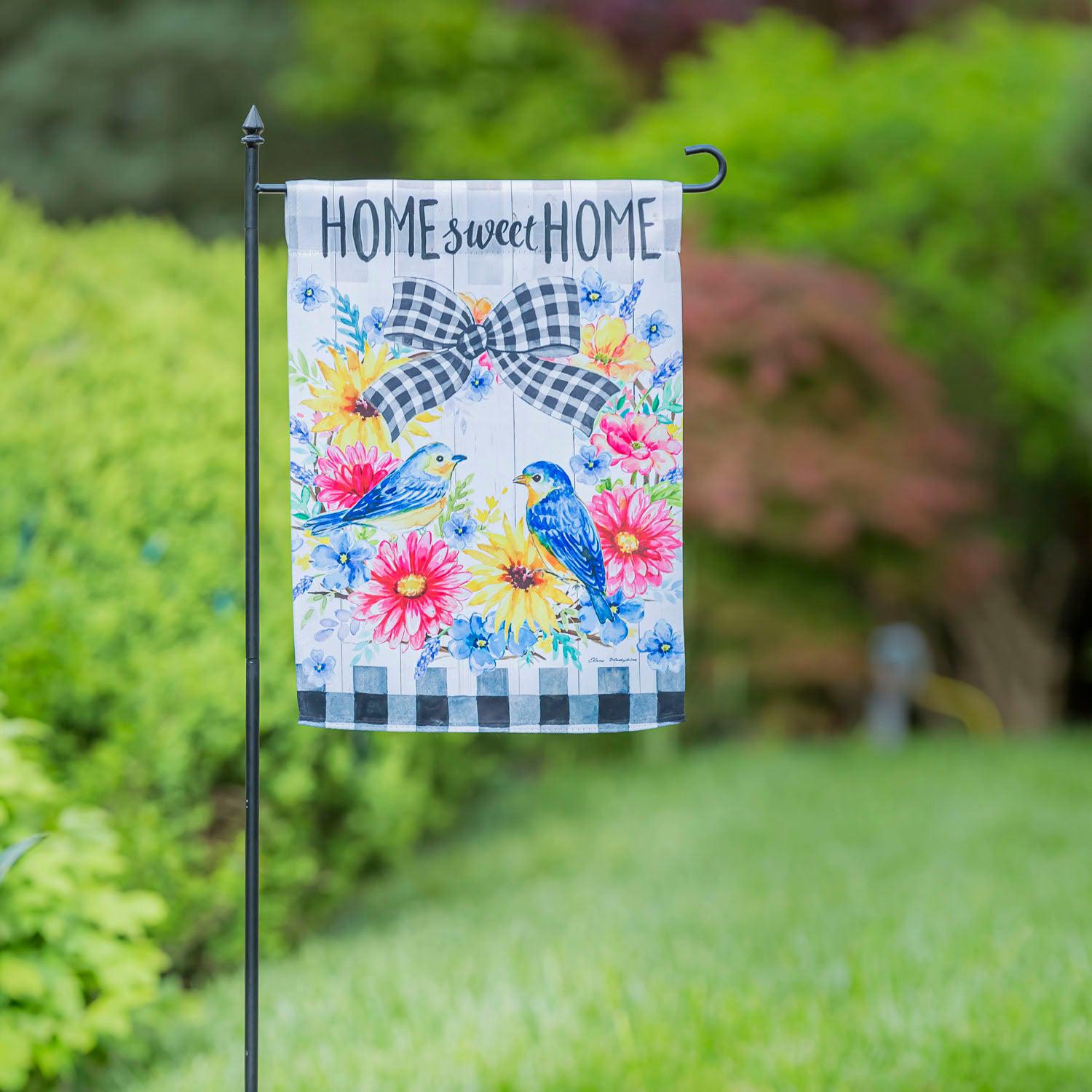 The Spring Bird Wreath garden flag features a spring floral wreath with a pair of bluebirds, black checked accents, and the words "Home Sweet Home".