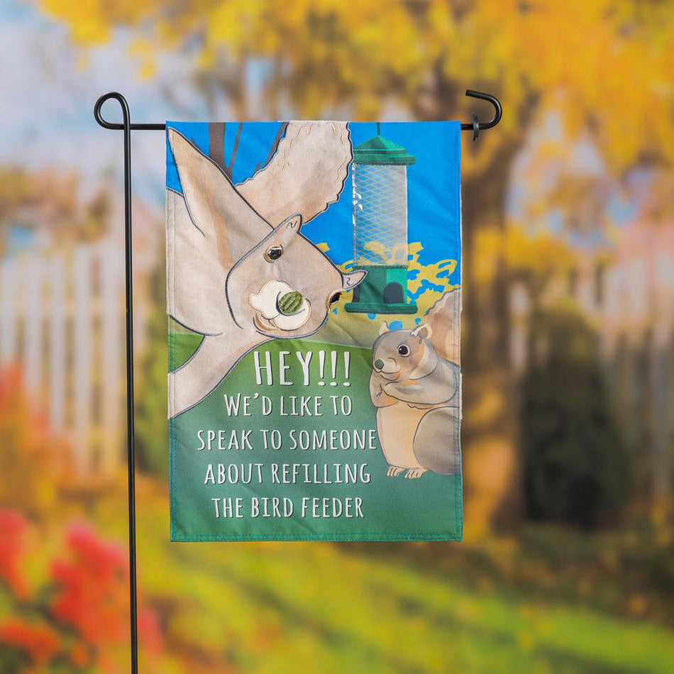 Our Squirrel Feeder garden flag features two squirrels appearing to look at you, an empty bird feeder, and the words "HEY!!! We'd like to speak to someone about refilling the bird feeder". 